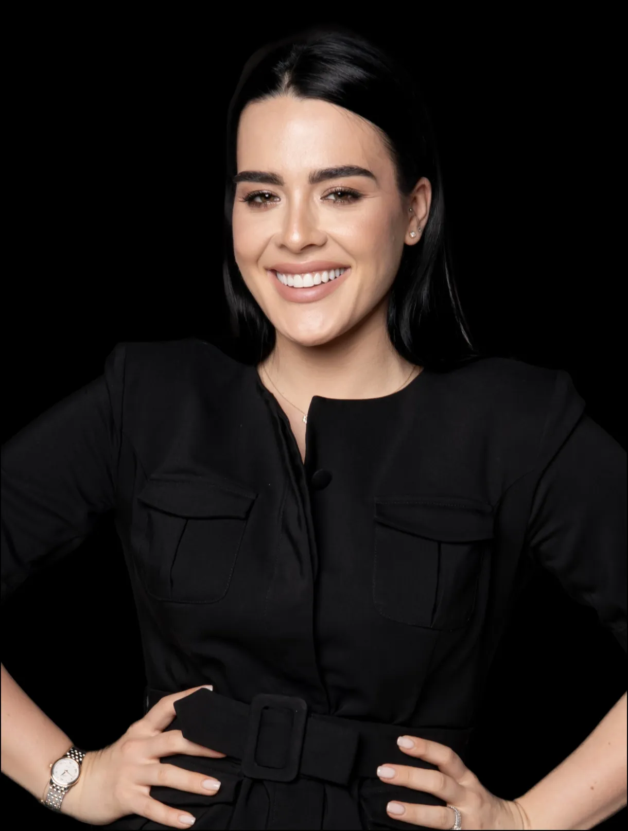 Kobieta o prostych, ciemnych włosach uśmiecha się pewnie. Ubrana w czarny top w paski z kieszeniami na piersi, pozuje na prostym czarnym tle, uosabiając elegancję często spotykaną u profesjonalistów z branży medycyny estetycznej we Wrocławiu.