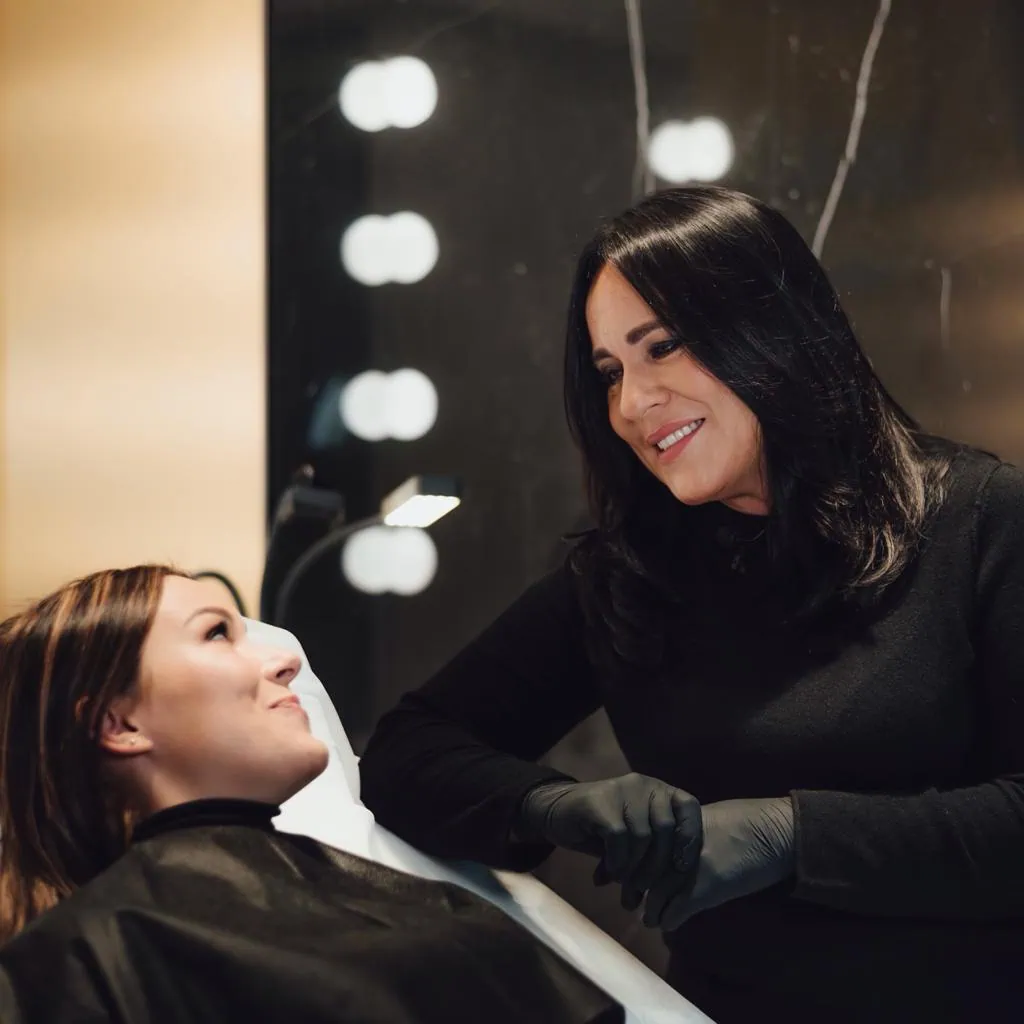 Kobieta w czarnych rękawiczkach uśmiecha się i rozmawia z kobietą leżącą na fotelu w salonie, obie zrelaksowane w dobrze oświetlonym pokoju z lustrem - zachęcająca scena typowa dla zabiegów medycyny estetycznej Wrocław lub botoks Wrocław.