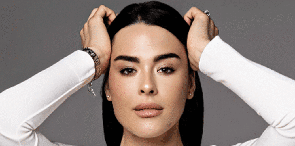 TERMOLIFTING CZY HIFU Osoba o prostych czarnych włosach, nieskazitelnej cerze i naturalnym makijażu pozuje z obiema rękami na głowie, prezentując promienne efekty medycyny estetycznej Wrocław, ubrana w białą bluzkę z długim rękawem i srebrną biżuterię na jednolitym szarym tle.