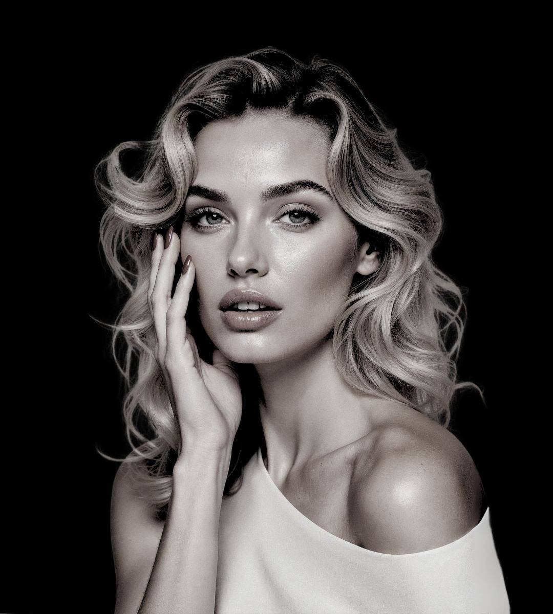 Kobieta o falujących blond włosach i jasnych oczach pozuje na czarnym tle, ubrana w biały top z odkrytymi ramionami. Delikatnie dotyka swojej twarzy, a jej pogodne spojrzenie odzwierciedla pewność siebie, która pochodzi z zabiegów botoks Wrocław.