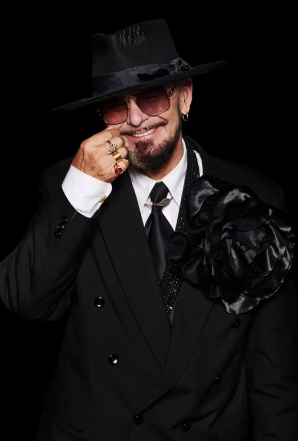 Osoba w czarnym garniturze, dużych czarnych kwiatowych szpilkach i czarnym kapeluszu uśmiecha się i dotyka twarzy. Z przyciemnianymi okularami przeciwsłonecznymi i zarostem na twarzy, uzupełnia się pierścionkami i kolczykami - emanując pewnością siebie jak klient medycyny estetycznej Wrocław.