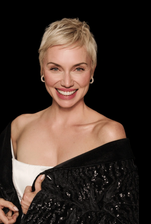 Uśmiechnięta kobieta o krótkich blond włosach, ubrana w kolczyki koła, biały top i czarną cekinową kurtkę przewieszoną przez ramiona, pozuje pewnie na prostym czarnym tle - promieniując wyrafinowanym pięknem medycyny estetycznej Wrocławia.