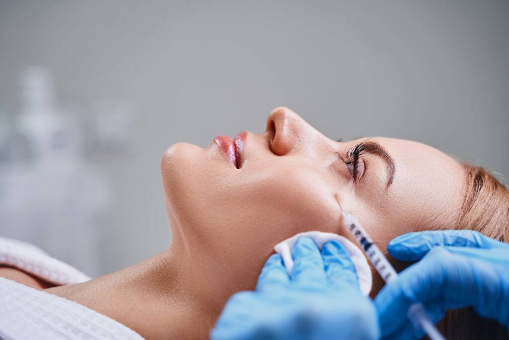A woman lies down at a klinika medycyny estetycznej, receiving a cosmetic injection near her cheekbone from a person in blue gloves, while her face is gently supported with a cotton pad.