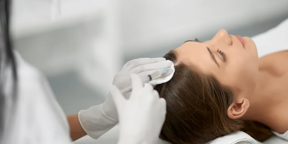 A woman lies on a treatment table as a gloved professional cleans her scalp with a cotton pad, preparing her for a hair or scalp treatment at a renomowana klinika medycyny estetycznej.