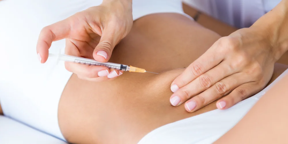 A person in white clothing injects a syringe into the skin of their lower abdomen, using one hand to pinch the skin—a scene often seen in klinika medycyny estetycznej or during medycyna estetyczna Wrocław procedures.