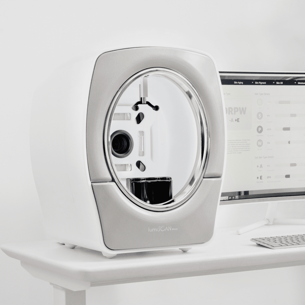 A modern, white, square-shaped medical imaging device with a circular opening sits on a white desk next to a computer monitor and keyboard in a bright, minimalist klinika medycyny estetycznej setting.