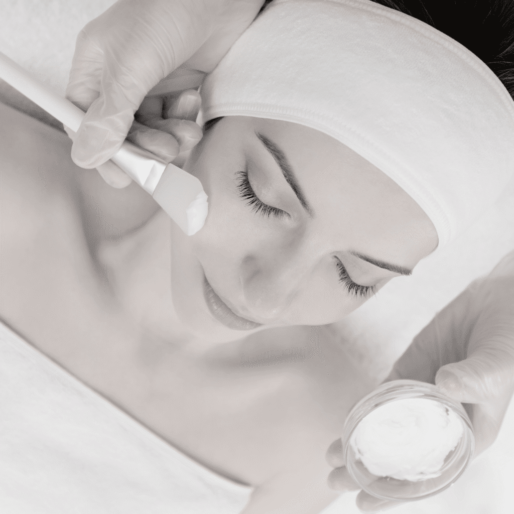 A woman with closed eyes and a headband is receiving a facial treatment at a klinika medycyny estetycznej, as a gloved hand applies a creamy mask to her cheek with a brush while another hand holds the small bowl of cream.