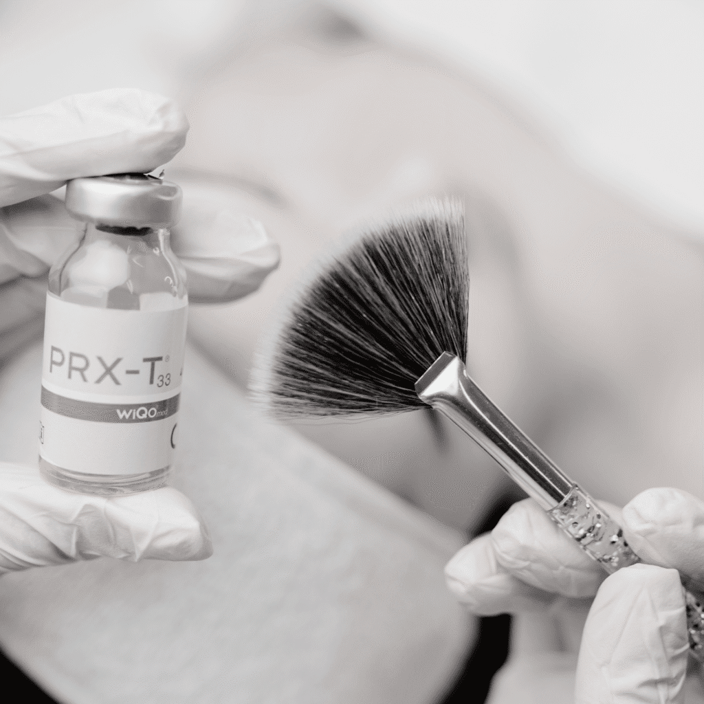A gloved hand holds a vial labeled PRX-T33 while another holds a fan brush, set against a blurred background at a klinika medycyny estetycznej, suggesting a professional skincare or cosmetic procedure.