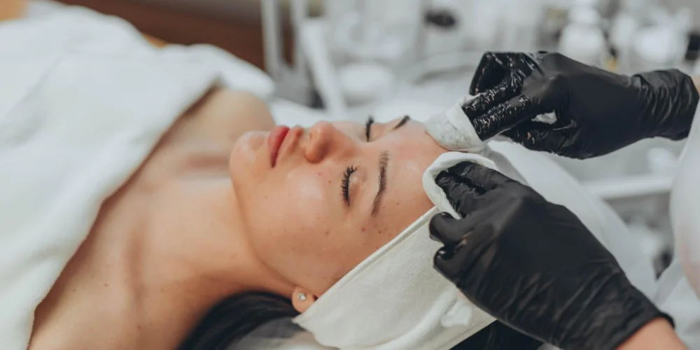 A person with closed eyes lies on a treatment bed, a towel wrapped around their head, as an esthetician at a klinika medycyny estetycznej gently cleanses their forehead with a cotton pad.