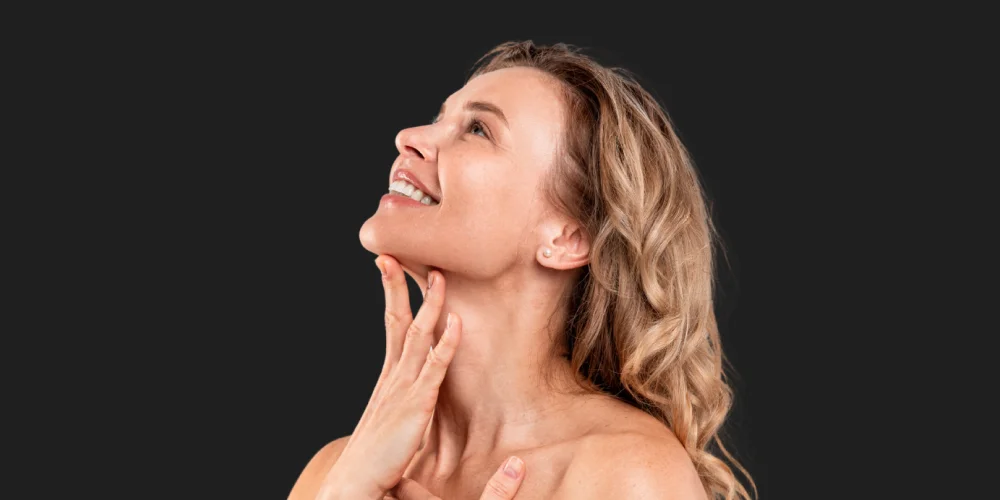 A smiling woman with wavy blonde hair looks upward, gently touching her neck with one hand. She appears relaxed and confident—radiating the results of medycyna estetyczna Wrocław. The background is plain black.