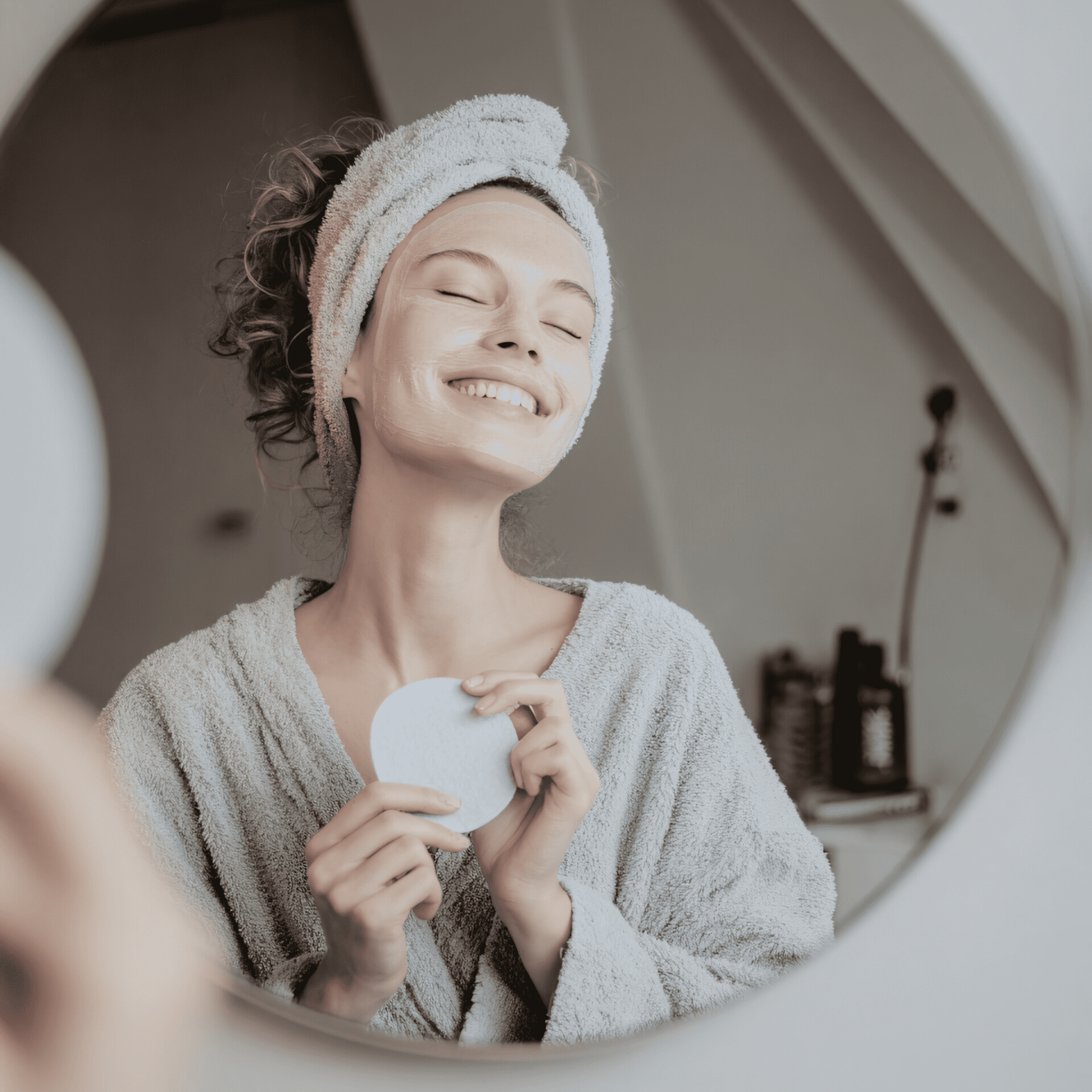 Jak przygotować skórę do zabiegów laserowych? A woman in a bathrobe and hair towel smiles with her eyes closed in front of a bathroom mirror, holding a cotton pad. She has a white facial mask on, enjoying a moment of self-care inspired by medycyna estetyczna Wrocław.