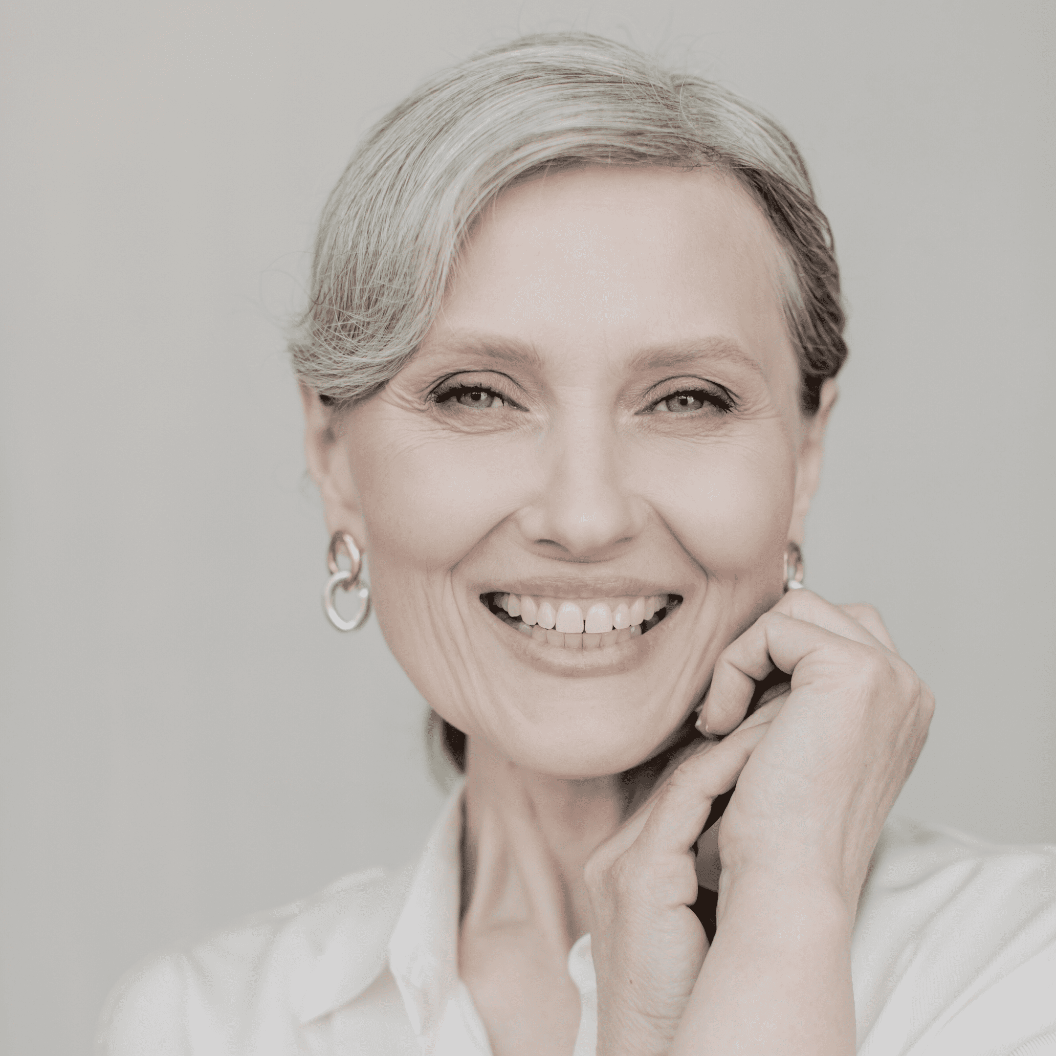 A smiling older woman with gray hair styled back, wearing hoop earrings and a white blouse, poses with one hand near her chin against a light background—radiating confidence and beauty after visiting klinika medycyny estetycznej.