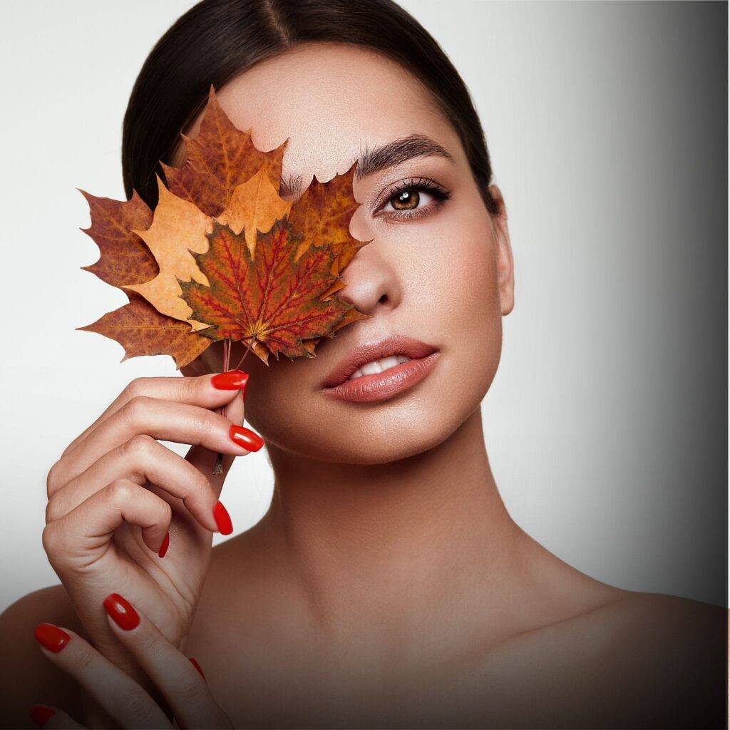 Woman with smooth skin and red nails holds several autumn leaves over one eye, looking at the camera against a neutral background—an image evoking the elegance of medycyna estetyczna Wrocław.
